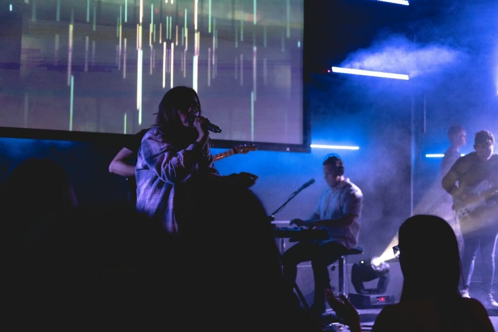 boy singing with microphone beside man playing guitar, near man playing electronic keyboard, and man playing bass guitar on stage