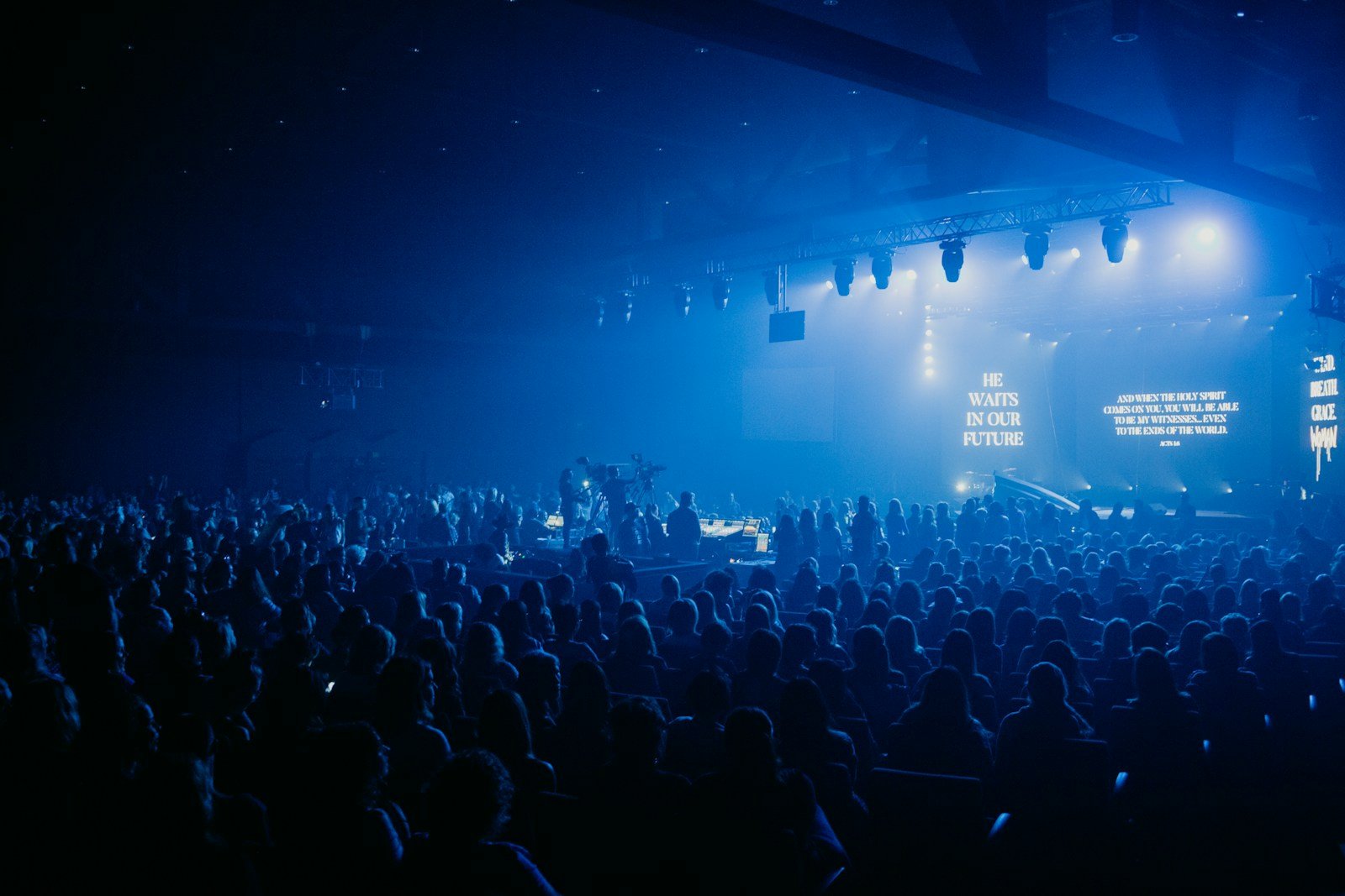 A large crowd watches a concert on a blue-lit stage.